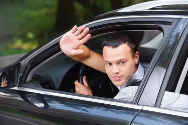 Portrait of young man driving car and greeting somebody with han