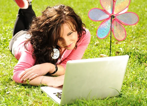 Happy young girl laying on green grass in nature and working on laptop