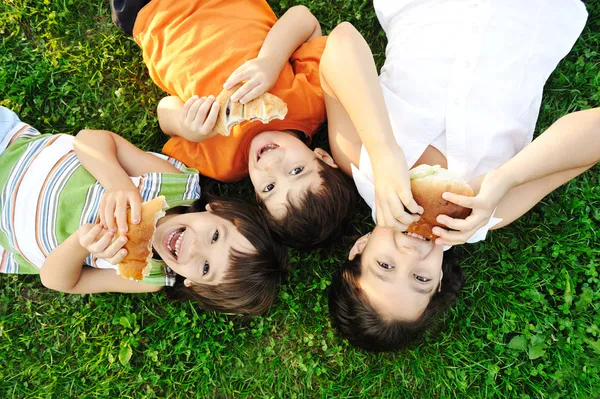 Three children laying on green grass on ground and eating sandwiches and sm