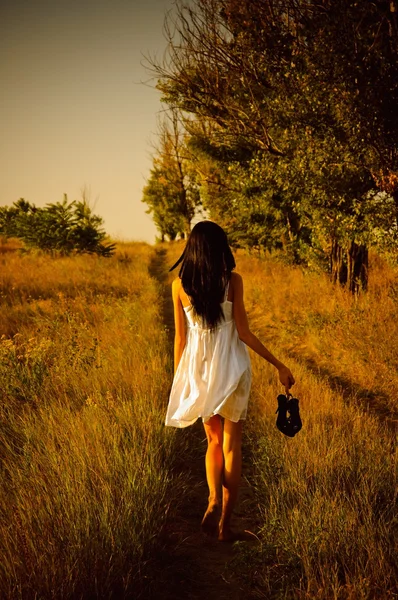 Barefoot girl in white dress with shoes in hand is on the field.