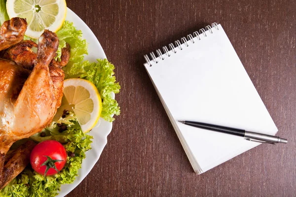 Restaurant table with roast chicken, notebook and pen — Stock Photo, Image
