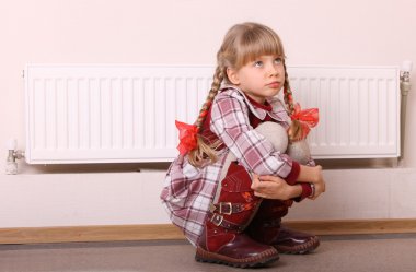 Sad girl sitting near heater. Children problem. clipart