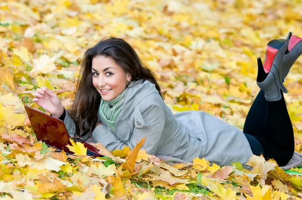Счастливая студентка лежит в осенних листьях с нетбуком — стоковое фото