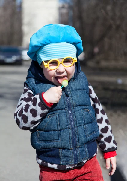 Little child with lollipop is standing outdoors. Funny glasses — Stock Photo, Image