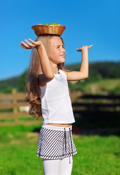 Niedliches kleines Mädchen mit langen blonden Haaren trägt Obstkorb auf dem Kopf — Stockfoto