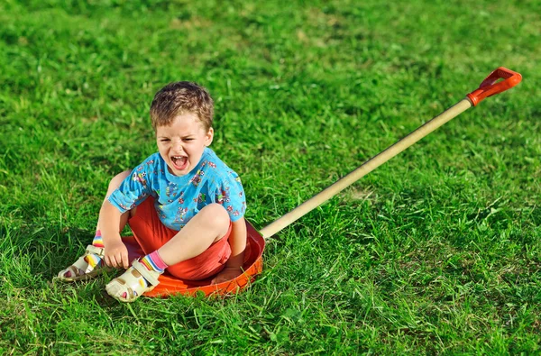 Маленький мальчик сидит на красной пластиковой лопате над зеленой газоном травы — стоковое фото