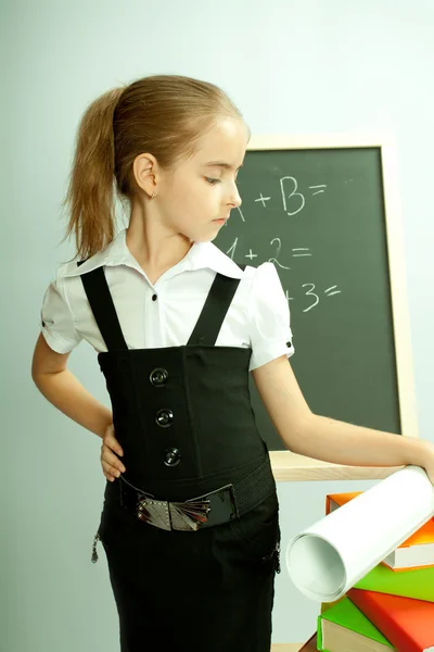School girl front blackboard with hand on stack of books. — Stock Photo, Image