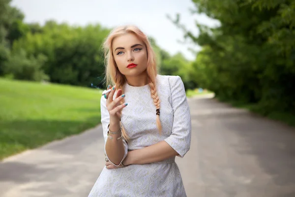 Fashionable blonde smoking a cigarette walks in park. — Stock Photo, Image