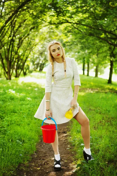 Fashionable young blonde poses in park avenue with a red bucket. — Stock Photo, Image