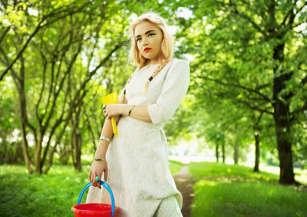 Modische junge Blondine posiert in Parkallee mit rotem Eimer. — Stockfoto