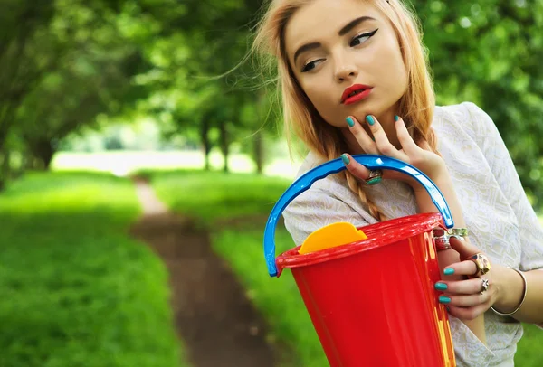Jovem loira na moda posa na avenida do parque com um balde vermelho . — Fotografia de Stock