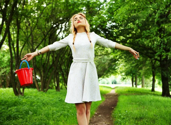 La giovane bionda alla moda posa in viale di parco con un secchio rosso . — Foto Stock
