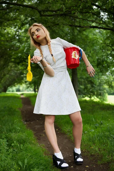 Fashionable young blonde poses in park avenue with a red bucket. — Stock Photo, Image