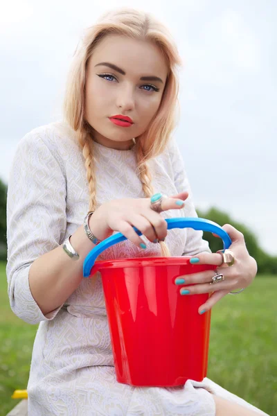 Stylish blonde with a red bucket on a green meadow. — Stock Photo, Image