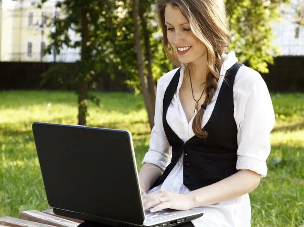 Fiatal, csinos nő ül a padon a parkban laptop — Stock Fotó