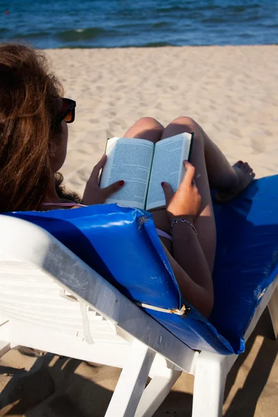 Vrouw liggen op een chaise lounge lezen van een boek op het strand — Stockfoto