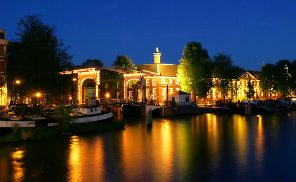 View on small bridge and illuminated houses on city canal (Amstel river) at evening in Amsterdam, Netherlands (Holland).