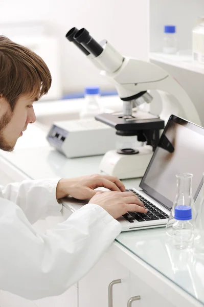 Young male researcher looking into a microscope and writing notes on laptop — Stock Photo, Image