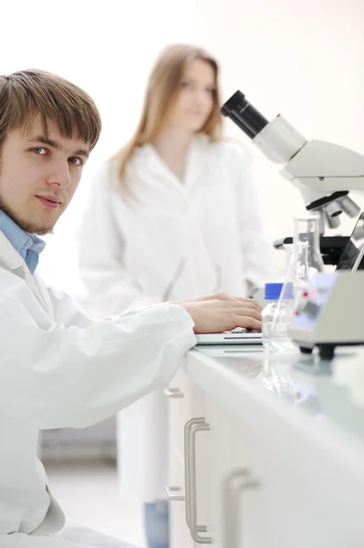 Young male researcher with a microscope and writing notes on laptop at labo — Stock Photo, Image