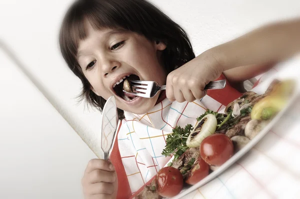 Lindo chico positivo con tenedor y cuchillo comiendo en la mesa del almuerzo —  Fotos de Stock