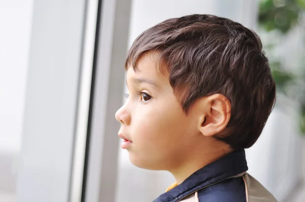 Porträtt av lilla ganska tankeväckande pojke tittar ut genom fönstret — Stockfoto