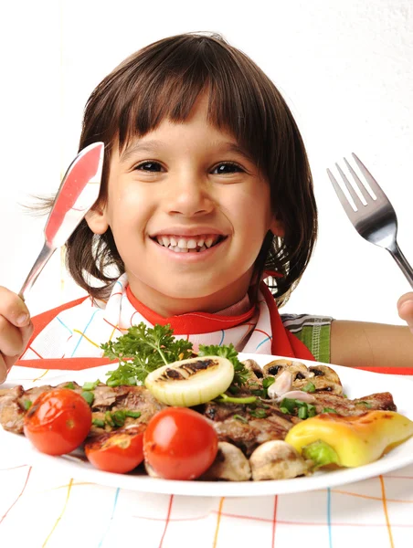 Söt positiva pojke med gaffel och kniv äta lunch tabellen — Stockfoto