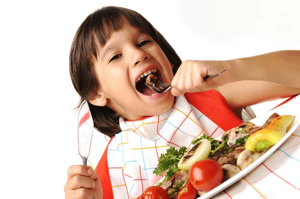 Симпатичный позитивный мальчик с вилкой и ножом ест на обеденном столе — стоковое фото