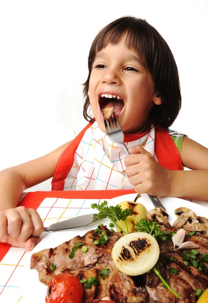 Bonito menino positivo com garfo e faca comendo na mesa de almoço — Fotografia de Stock