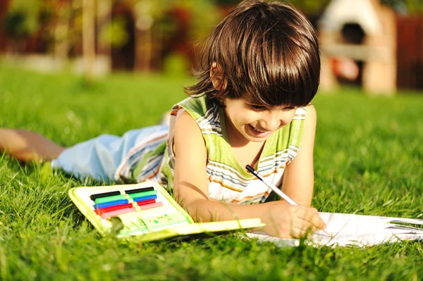 Young boy outdoors on the grass reading a book, writting and drawing — Stock Photo, Image