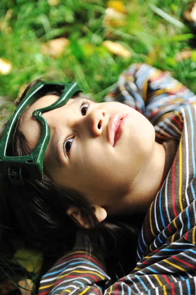 Mignon garçon positif avec des lunettes reposant sur le sol d'herbe verte et levant les yeux — Photo