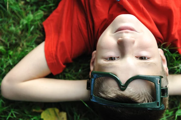 Cute positive boy with glasses laying on green grass ground and looking up — Stock Photo, Image