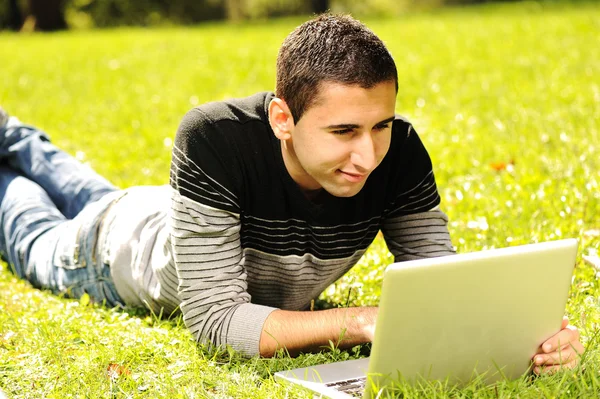 Feliz joven acostado en la hierba verde en la naturaleza y trabajando en el ordenador portátil —  Fotos de Stock