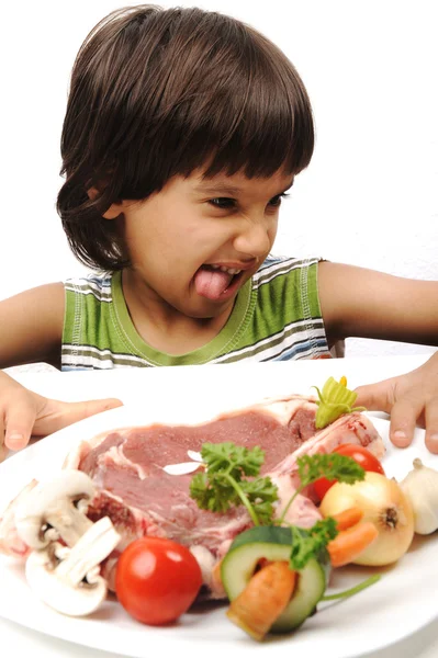Cute positive boy and raw meat and vegetables in plate reading for cooking — Stock Photo, Image
