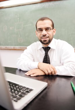 Happy positive teacher with laptop on desk in classroom clipart