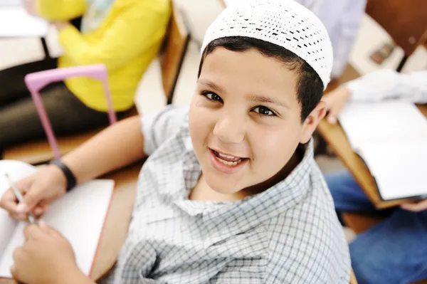 Niño muy positivo con sombrero blanco pequeño sentado en el escritorio en el aula y smi —  Fotos de Stock