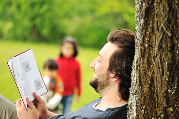 Fiatal laza apa olvasó a könyvet, és ezen a környéken: játszó gyermekek — Stock Fotó