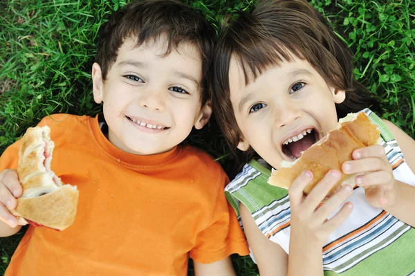 Dos chicos lindos tendidos en el suelo en la naturaleza y comiendo felizmente alimentos saludables Imagen de archivo