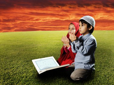 Two Muslims children sitting on meadow in Ramadan and reading Koran and pra