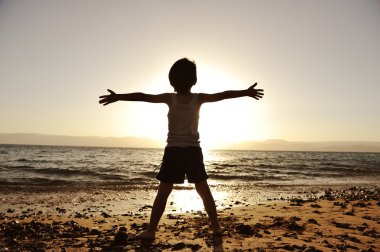 Silhouette of child on the beach, holding his hands up, towards the sun clipart