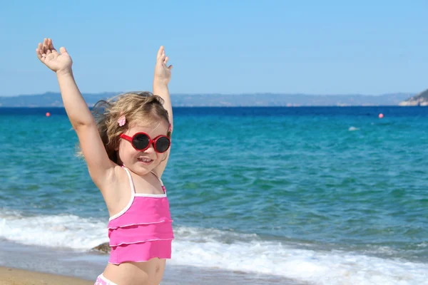 Ragazzina felice con occhiali da sole e mani in alto sulla spiaggia — Foto Stock