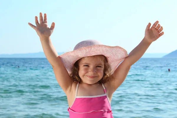 Счастливая маленькая девочка в соломенной шляпе и с поднятыми руками на пляже — стоковое фото
