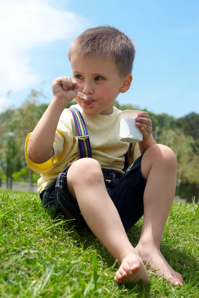 Маленький хлопчик апетитно їсть йогурт, сидячи на траві — стокове фото