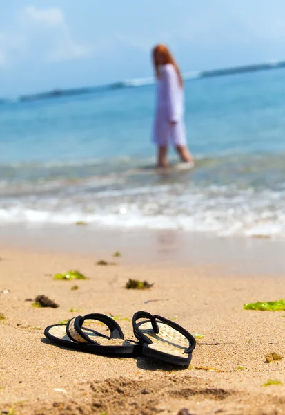 Zapatillas de playa en la arena y chica en el océano fuera de foco — Foto de Stock