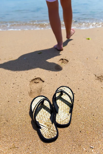 Las zapatillas de playa sobre la arena, y los pies femeninos al borde del mar —  Fotos de Stock