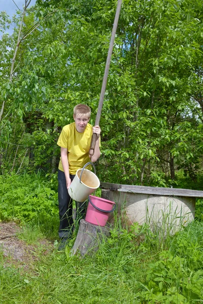 O menino, o adolescente, derrama água em um balde de um poço — Fotografia de Stock