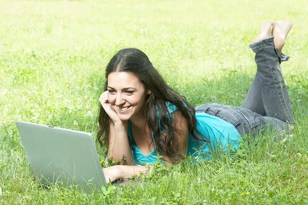 Retrato de uma linda jovem mulher deitada na grama no parque usando um lapt — Fotografia de Stock