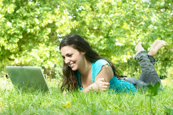 Porträt einer niedlichen jungen Frau, die mit einem Laptop auf dem Rasen im Park liegt — Stockfoto