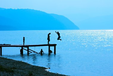 Silhouettes of kids who jump off dock on the lake clipart