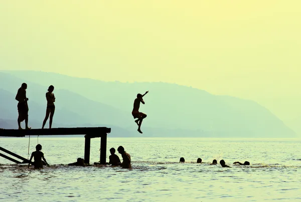 Silhouettes of kids who jump off dock on the lake — Stock Photo, Image