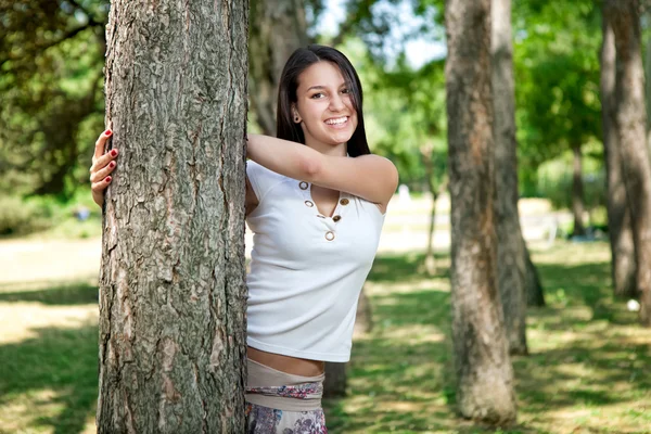 Gelukkig jong meisje verschuilt zich achter een boom in het park — Stockfoto